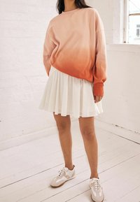 Oversized bluza w różowo-pomarańczowym gradiencie, biała plisowana spódnica i jasne sneakersy. Charakteryzuje się miękką teksturą i luźnym krojem.