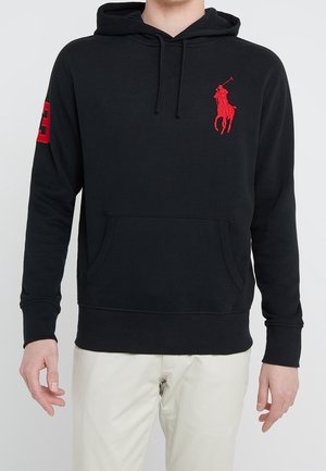 Mężczyzna w czarnej bluzie z kapturem z czerwonym logo gracza polo na piersi i czerwonym numerem 3 na rękawie, w połączeniu z kremowymi spodniami.