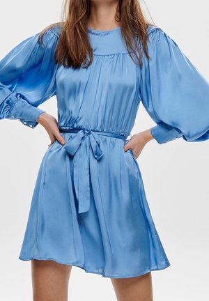 Kobieta w jasnoniebieskiej satynowej sukience z długimi bufkami i wiązanym paskiem w talii, stojąca z rękami na biodrach.