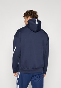 Marinblauer Hoodie aus weichem Material mit großer Kapuze, seitlichen weißen Akzenten und einem Etikett auf der Rückseite. Kombiniert mit passenden blauen Hosen.