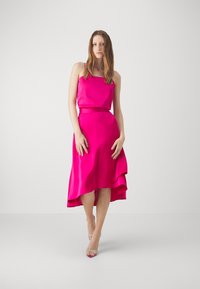Fuchsia satin dress with a fitted bodice, flowing asymmetrical hem, and thin straps. Paired with clear heeled sandals.