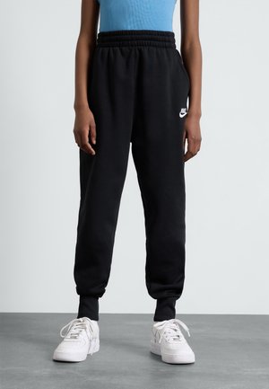 Personne portant un pantalon de survêtement Nike noir avec ceinture et poches élastiques, associé à des baskets blanches et un haut bleu clair.