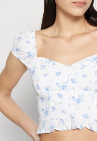 Kvinna som bär en vit kortärmad blus med blå blommönster, puffärmar, framknappar och volangkant, visad från halsen till midjan.