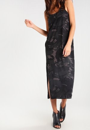 Femme portant une robe midi sans manches à motif camouflage noir et gris avec une fente latérale et des bottines noires à bouts ouverts avec fermetures éclair à l'avant.