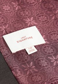Tissu bordeaux à motifs floraux, avec une étiquette blanche indiquant "NEXT SIGNATURE" et la taille "42R", signalant une origine du Vietnam.