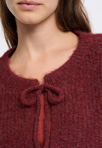 Maglione lavorato a maglia color mogano con scollatura rotonda, tessuto testurizzato e chiusura con lacci al colletto, indossato sopra un indumento rosso.