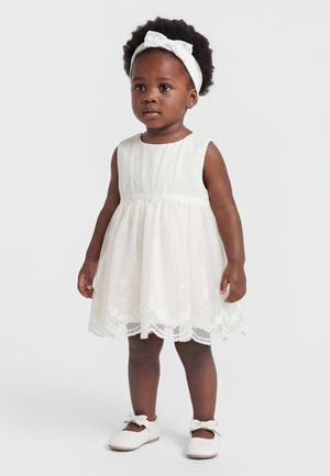 Petite fille en bas âge aux cheveux bouclés foncés portant une robe blanche sans manches, un bandeau assorti avec un nœud, et des chaussures blanches avec des nœuds, debout sur fond blanc.