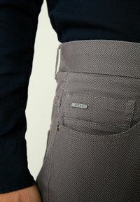 Nærbillede af en person iført teksturerede grå bukser med et lille metalskilt mærket "NEXT" på baglommen og en mørk langærmet top.
