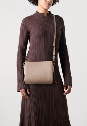 Borsa a tracolla in pelle beige con una texture liscia, hardware oro e tracolla regolabile. Indossata su un vestito marrone a coste.