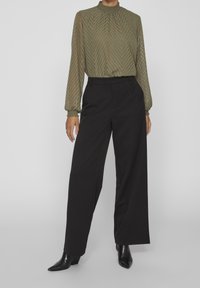 Blouse en vert olive, en voile, avec un motif chevron, col haut et détails froncés, assortie à un pantalon noir à jambes larges et des bottines noires à bout pointu.