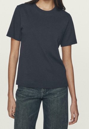 Kvinde iført en ensfarvet mørk navy kortærmet T-shirt med rund hals og blå jeans, stående mod en ensfarvet lys baggrund.