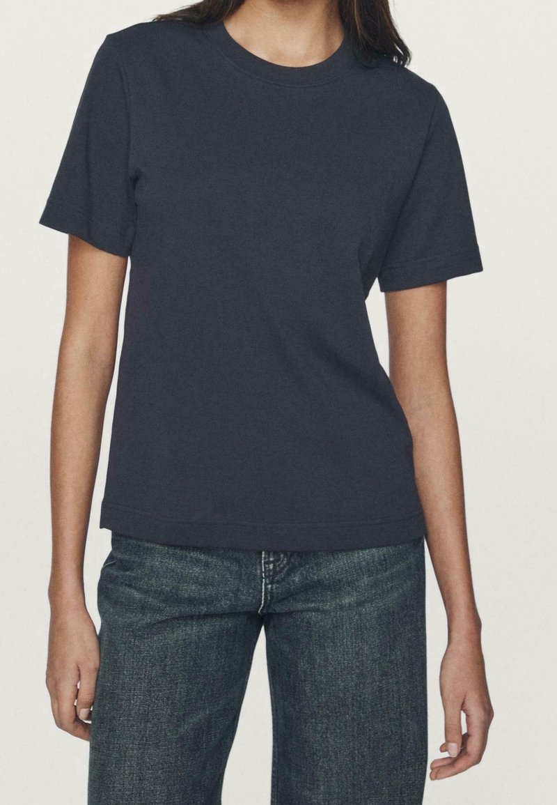 Femme portant un T-shirt uni bleu marine foncé à manches courtes et col rond, et un jean bleu, debout devant un fond clair uni.