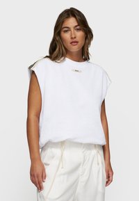 Top sin mangas blanco con una tela texturizada, que presenta un escote redondo y una pequeña etiqueta con logo. Combinado con pantalones de tiro medio de color claro con cordón.