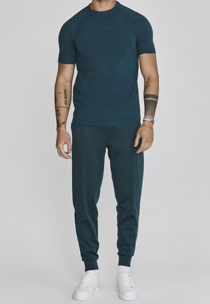 Homme portant un t-shirt bleu sarcelle foncé ajusté et un pantalon de jogging assorti, des baskets blanches, des tatouages visibles sur les bras et des accessoires au poignet, debout devant un fond uni.