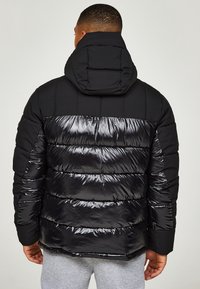 Svart pufferjacka med blanka detaljer och en strukturerad huva. Har en vertikalt sydd design och grått foder.