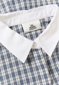 Camisa de cuello con un patrón a cuadros en tonos azules y blancos, con un cuello blanco sólido y una etiqueta de logo cosida en el interior.