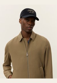 Giacca beige con zip, colletto e tasche laterali, indossata sopra una camicia blu a fantasia; completata da un cappellino nero con testo dorato.