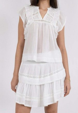 Vrouw in een witte mouwloze jurk met volants en kanten details, met een V-hals en een gelaagde rok.