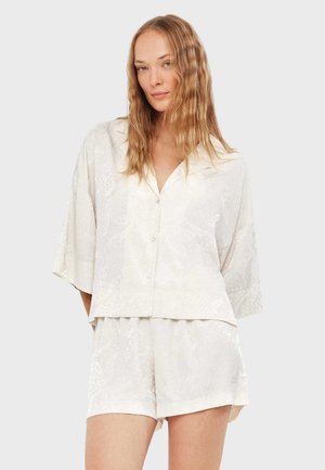 Mujer con cabello rubio ondulado, vestida con una camisa blanca suelta de manga corta con botones y pantalones cortos blancos a juego, sobre un fondo liso.