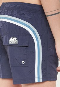 Pantaloni da bagno navy con tasca, vita elasticizzata e strisce laterali bianche e azzurre. Tessuto testurizzato con dettaglio del marchio.