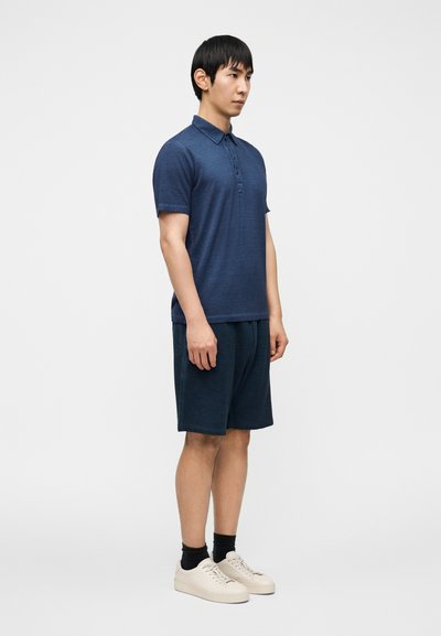 Μπλε σκούρο μπλουζάκι πόλο με κοντά μανίκια, με δύο κουμπιά στο ντεκολτέ, συνδυασμένο με σκούρα σορτς. Το μοντέλο φοράει ανοιχτόχρωμα αθλητικά παπούτσια και κάλτσες μέχρι τον αστράγαλο.