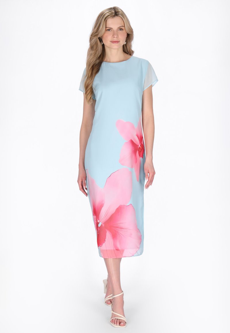 Žena v modrozelených midi šatech s velkými růžovými květinovými vzory, stojící v uvolněné pozici a obutá v bílé sandály na podpatku.