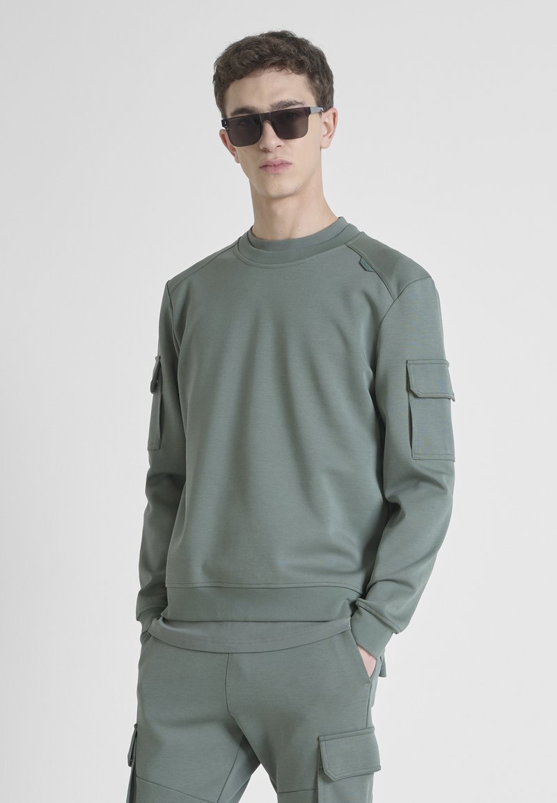Felpa grigio-verde con colletto rotondo, maniche lunghe e due tasche a lato. Texture liscia, stile aderente e dettagli unici sulle spalle.