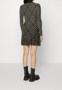 Femme portant une robe mini à manches longues noire et blanche à motifs et des bottines noires épaisses, se tenant devant un fond blanc uni.