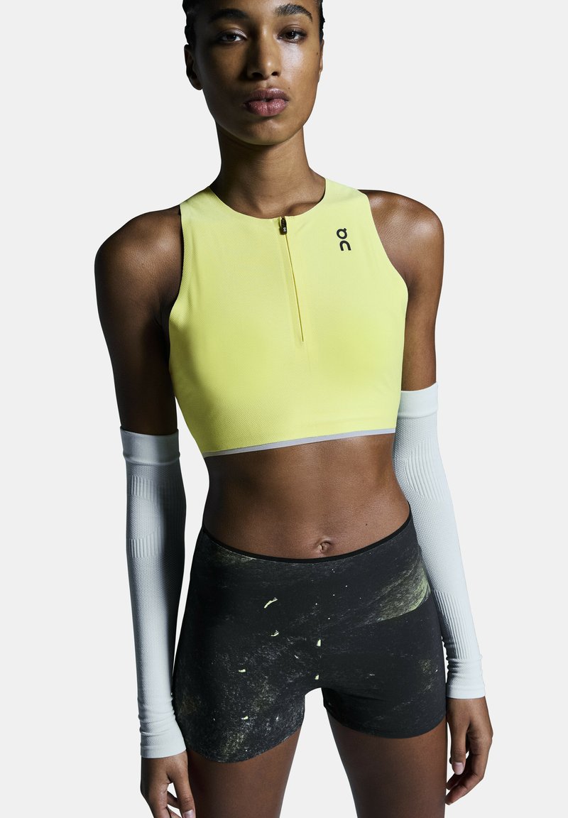 Femme portant un crop top jaune sans manches avec une petite fermeture éclair et un short noir à motifs, avec des manches blanches aux bras.