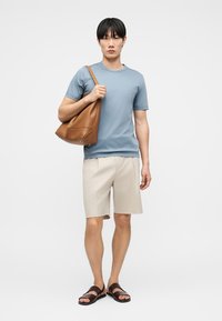 Άνδρας που φορά ανοιχτό μπλε κοντομάνικο πουκάμισο, μπεζ σορτς, σκούρα καφέ σανδάλια, κρατώντας μεγάλη καφέ δερμάτινη τσάντα ώμου.