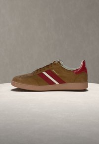 Καφέ σουέντ αθλητικό παπούτσι με κόκκινες και λευκές ρίγες στο πλάι, κόκκινη ταμπέλα στη φτέρνα και λογότυπο "Bally" στο πλάι και στη φτέρνα, τοποθετημένο σε επίπεδη επιφάνεια.