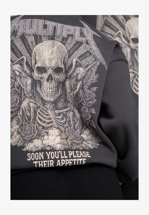 Zwarte trui met een prominent grafisch ontwerp van een schedel, rozen en skeletten, met metalen accenten en tekst die luidt: "BINNENKORT BEVREDIG JE HUN HONGER."