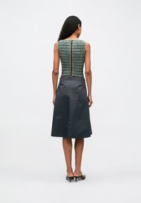 Ujjatlan, olívazöld steppelt felső hátsó cipzárral; sima anyagból készült, bővülő fekete szoknya; a modell fekete magassarkú cipőben áll.