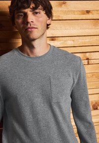 Camicia grigia a maniche lunghe in tessuto testurizzato con taschino sul petto, dotata di scollatura rotonda e vestibilità comoda, su uno sfondo di legno.