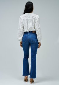Person facing away, wearing a white lace blouse and blue flared jeans. Standing on a plain background, showcasing the outfit's details.