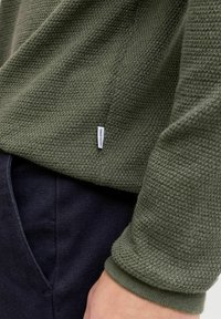 Close-up of a green textured knit sweater with a white Jack & Jones label, worn with dark navy pants and a visible hand resting nearby.