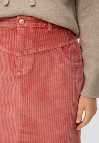 Pull beige rentré dans une jupe en velours côtelé rose taille haute avec une poche avant et un bouton, main reposant à côté de la jupe.