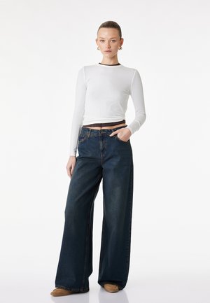 Vrouw staand, gekleed in een wit shirt met lange mouwen, hoge taille wijde donkerblauwe jeans en beige schoenen, met één hand in de zak tegen een witte achtergrond.