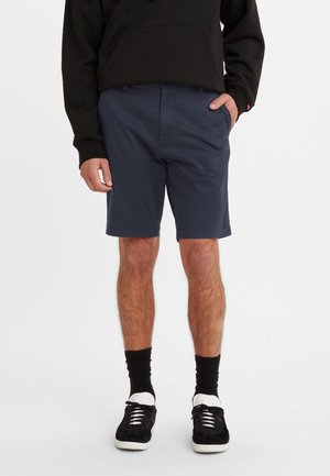 Pantalones cortos azul marino de algodón, con un corte recto y bolsillos laterales, llevados con una sudadera negra y calcetines negros, complementados con zapatillas negras.