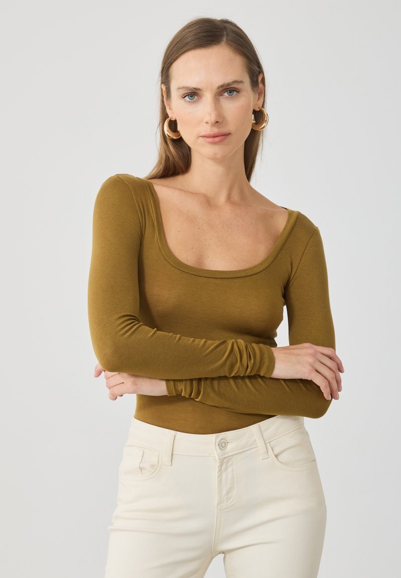 Top de manga longa, com decote em forma de barco, na cor verde oliva, feito de tecido suave. Apresenta um design ajustado e mangas texturizadas. Combinado com calças brancas.