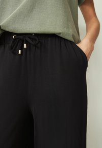 Sorte snørede bukser med elastisk talje; har guld-tone hardware. Båret med en grøn overdel, der viser en afslappet pasform og sidelommer.