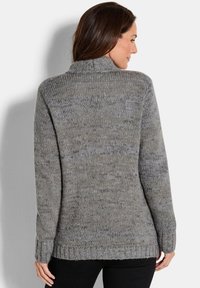 Frau in einem grauen gestrickten Pullover mit langen Ärmeln, die mit dem Rücken zu einer schlichten, hellen Hintergrund steht.