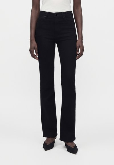Personne portant un pantalon noir taille haute à jambes droites avec un bouton et une fermeture éclair, associée à des talons noirs à bout carré et un haut blanc.