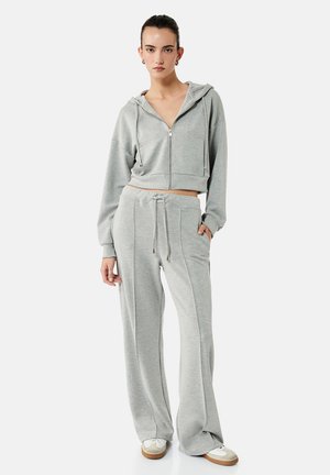 Junge Frau steht und trägt einen hellgrauen, kurz geschnittenen Kapuzenpullover mit Reißverschluss und passende weit geschnittene Sweatpants mit weißen Turnschuhen vor weißem Hintergrund.