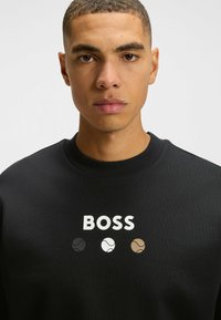 Sudadera negra con cuello redondo y un logo blanco de "BOSS" sobre tres pequeños diseños de pelotas de tenis en colores negro, blanco y marrón.