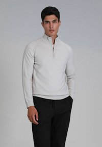 Hellgrauer Quarter-Zip-Pullover mit langen Ärmeln und gerippten Bündchen, kombiniert mit schwarzen Jogginghosen mit Kordelzug. Glatter Stoff mit taillierter Passform.