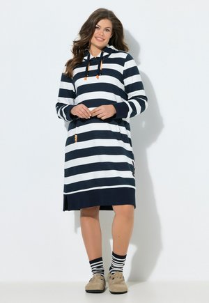 Kvinde iført navyblå og hvide stribede hættetrøjekjole, stående med hænderne foldet, kombineret med matchende stribede sokker og beige sko.