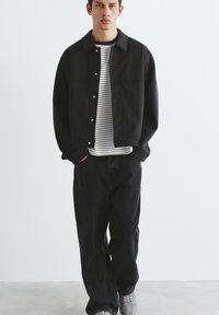 Jonge man staand met handen in de zakken, draag een zwart jack, zwarte broek, gestreept wit en zwart overhemd en grijze sneakers op een effen achtergrond.