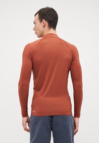 Camiseta atlética de manga larga en color óxido, con una tela suave y elástica y costuras planas para mayor comodidad. Diseño simple sin logotipos visibles.
