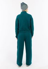 Persona in piedi di spalle, che indossa un sudario e pantaloni color teal con un berretto a maglia nero e bianco a strisce, su uno sfondo bianco.